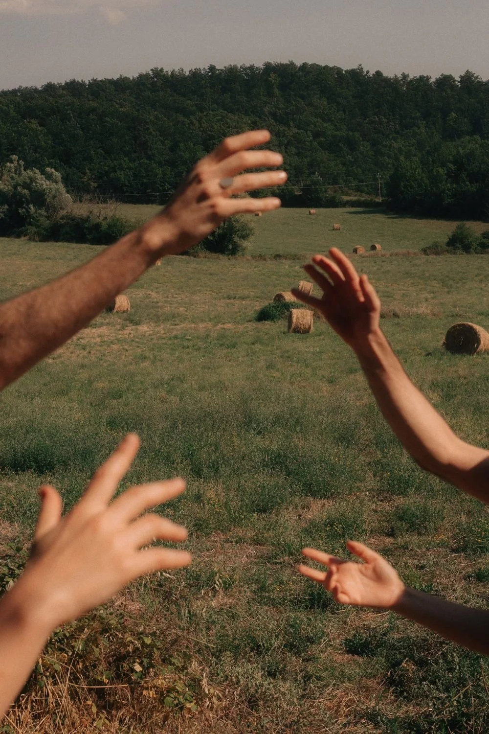 Three people reaching out their hands toward each other across a grassy field with hay bales and a tree-covered hill in the background.