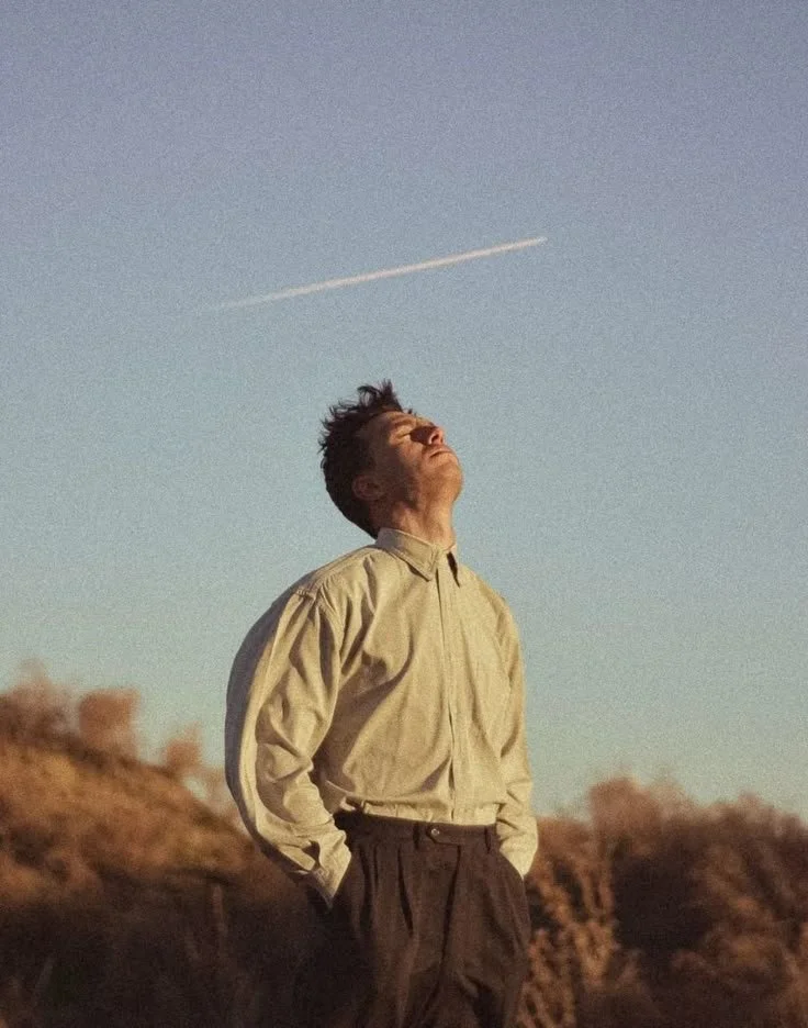 A man standing outdoors in a field, looking up with eyes closed as the sun shines on his face, with a clear sky and a jet trail above
