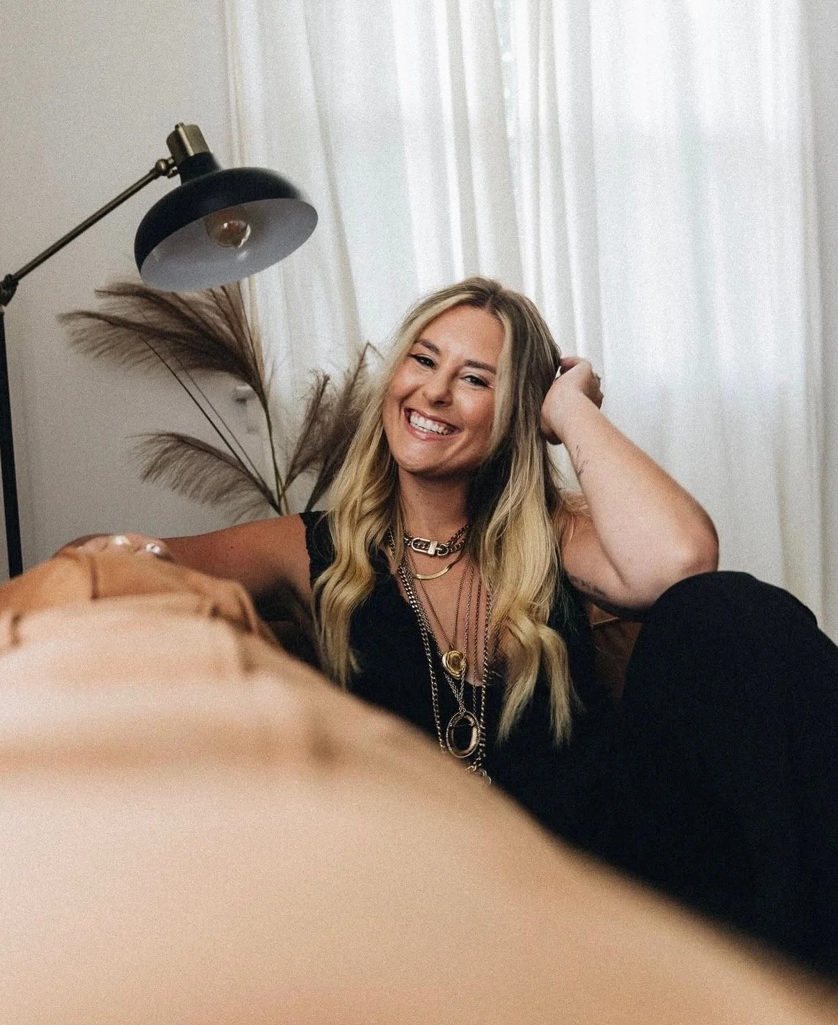 A woman with long blonde hair smiling and posing indoors, wearing layered necklaces and a black sleeveless top.