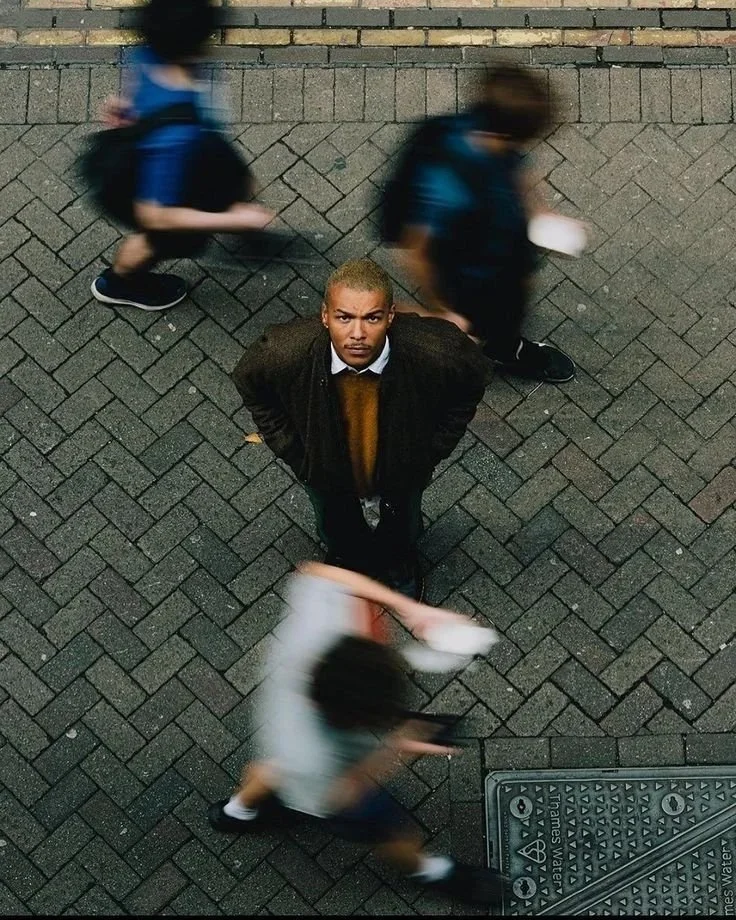 From above, a man with bleached blonde hair stands on a brick sidewalk looking up at the camera, surrounded by blurred pedestrians walking by.