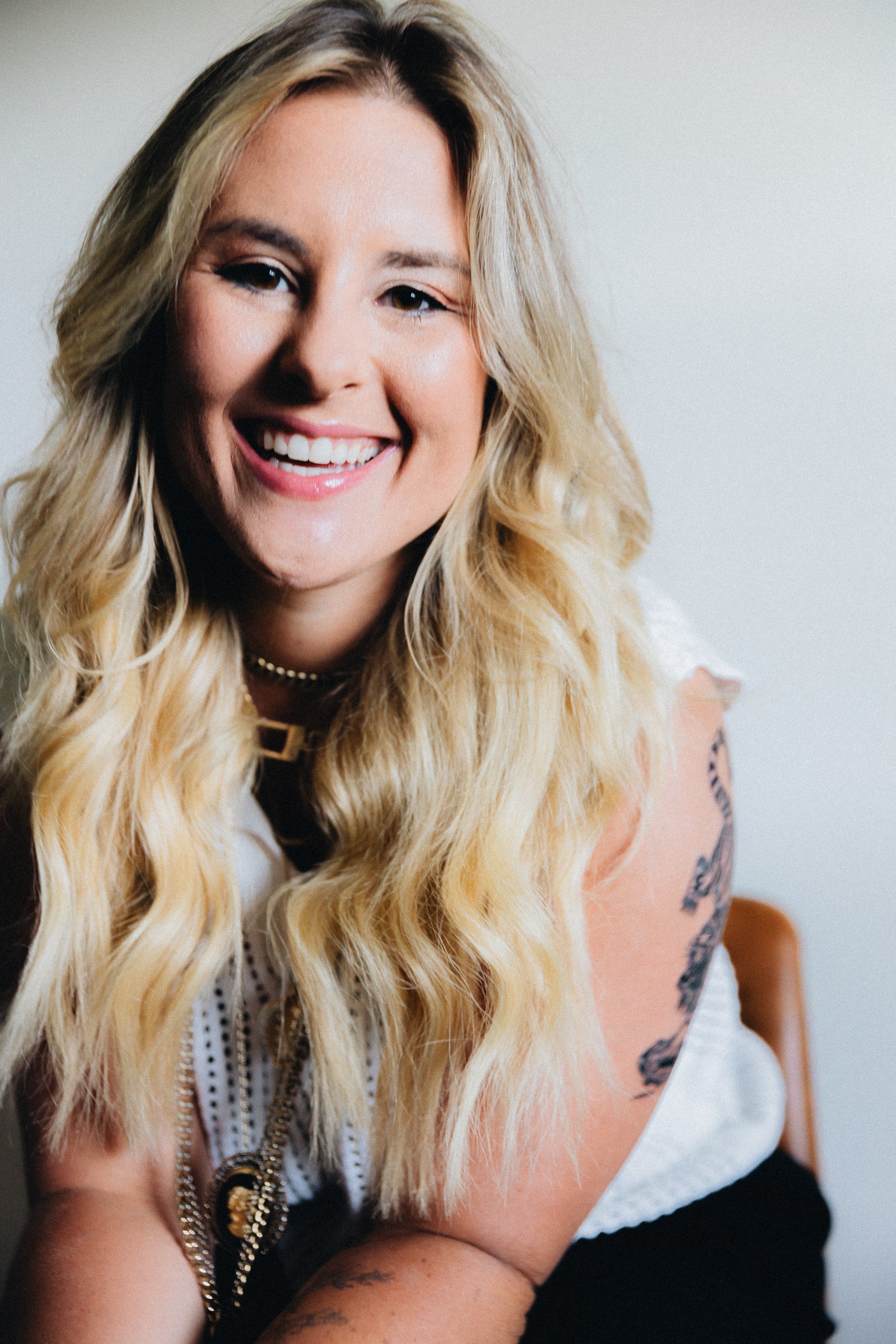 A woman with long blonde wavy hair, smiling, wearing a pearl necklace, layered chains, and a white top, with tattoos on her arms, sitting on a chair against a plain background.