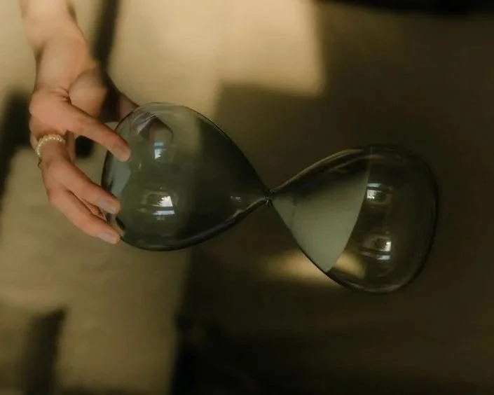 A hand holding a black hourglass with transparent glass bulbs on a beige background.