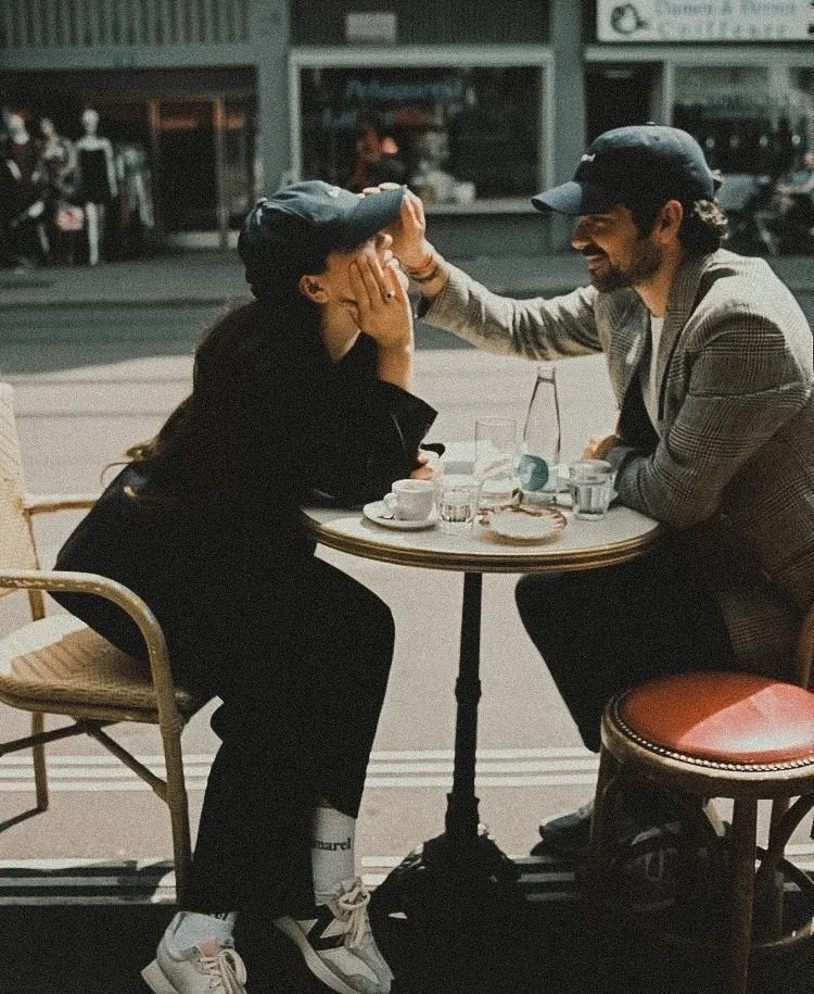 A couple sits at an outdoor cafe table, with the man gently touching the woman's face as they share a joyful moment.