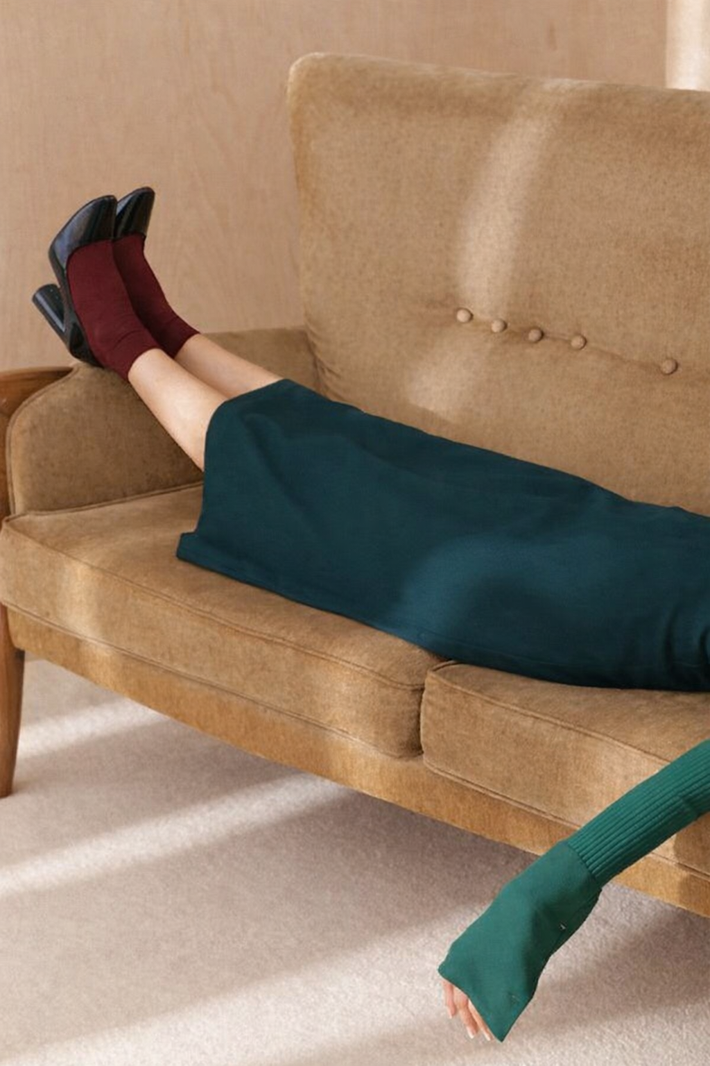 Person lying on a vintage brown couch with legs up, wearing red socks and black high heels, green skirt, and green long-sleeve top, in a room with beige carpet and wooden walls.