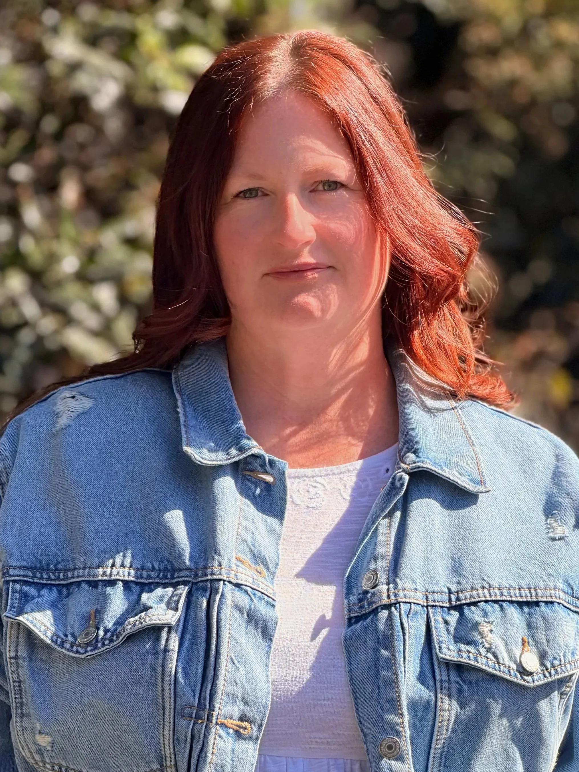 A woman with red hair wearing a denim jacket over a white top standing outdoors with sunlight and blurred greenery in the background.