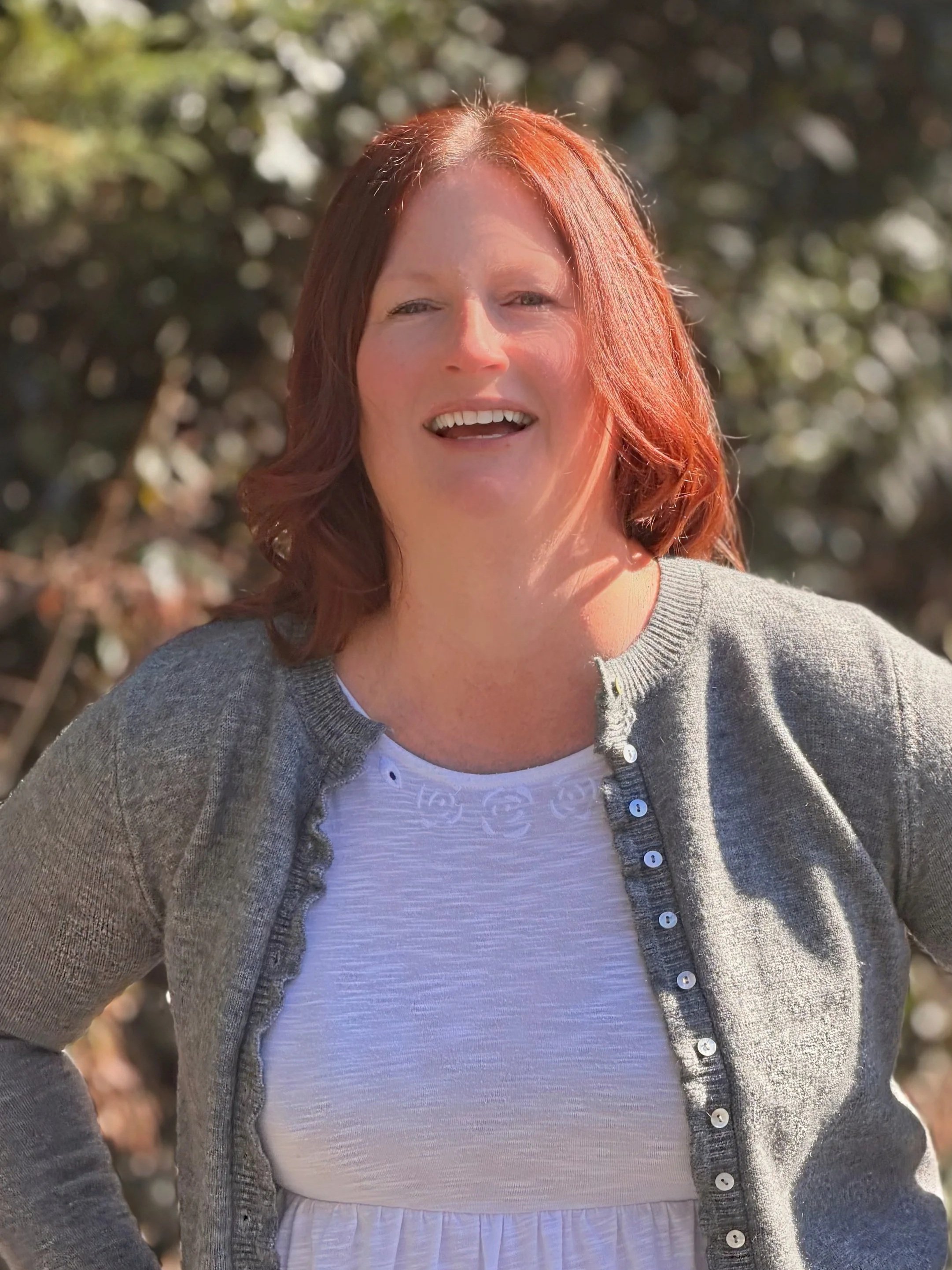 A woman with red hair smiling outdoors, wearing a light-colored top and a gray cardigan, with trees blurred in the background.