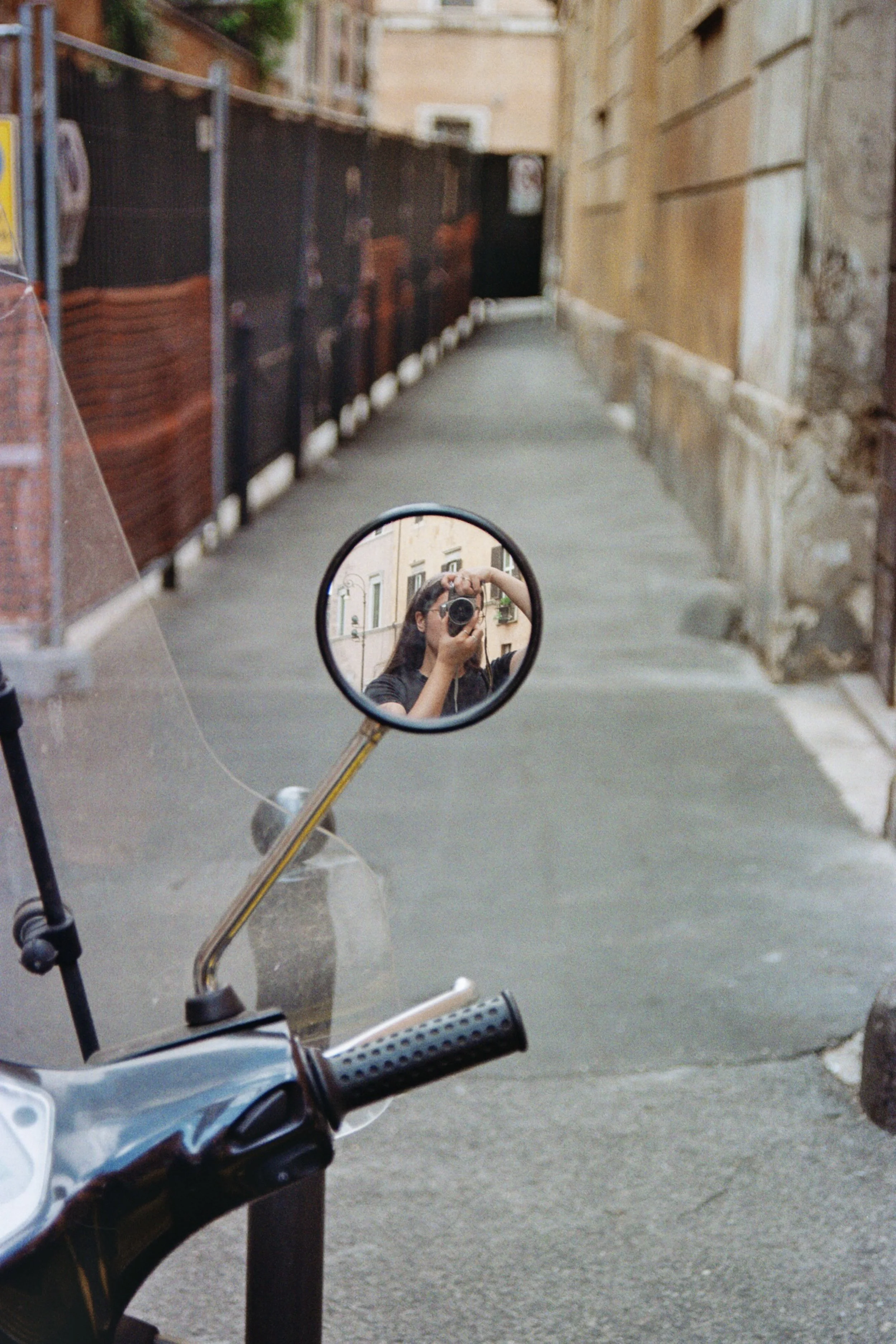Reflexo de uma pessoa tirando uma foto de si mesma com uma câmera na fechadura de um espelho de moto, visto em um espelho retrovisor de moto em uma rua estreita com paredes de edifícios antigos.