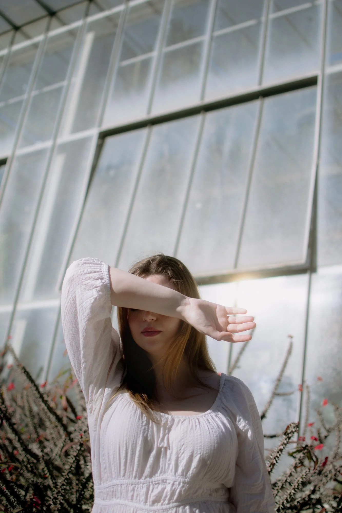 Mulher vestindo blusa branca, com cabelo castanho, cobre parcialmente o rosto com o braço, ao fundo vidro de uma estufa ou jardim de vidro e plantas com espinhos.