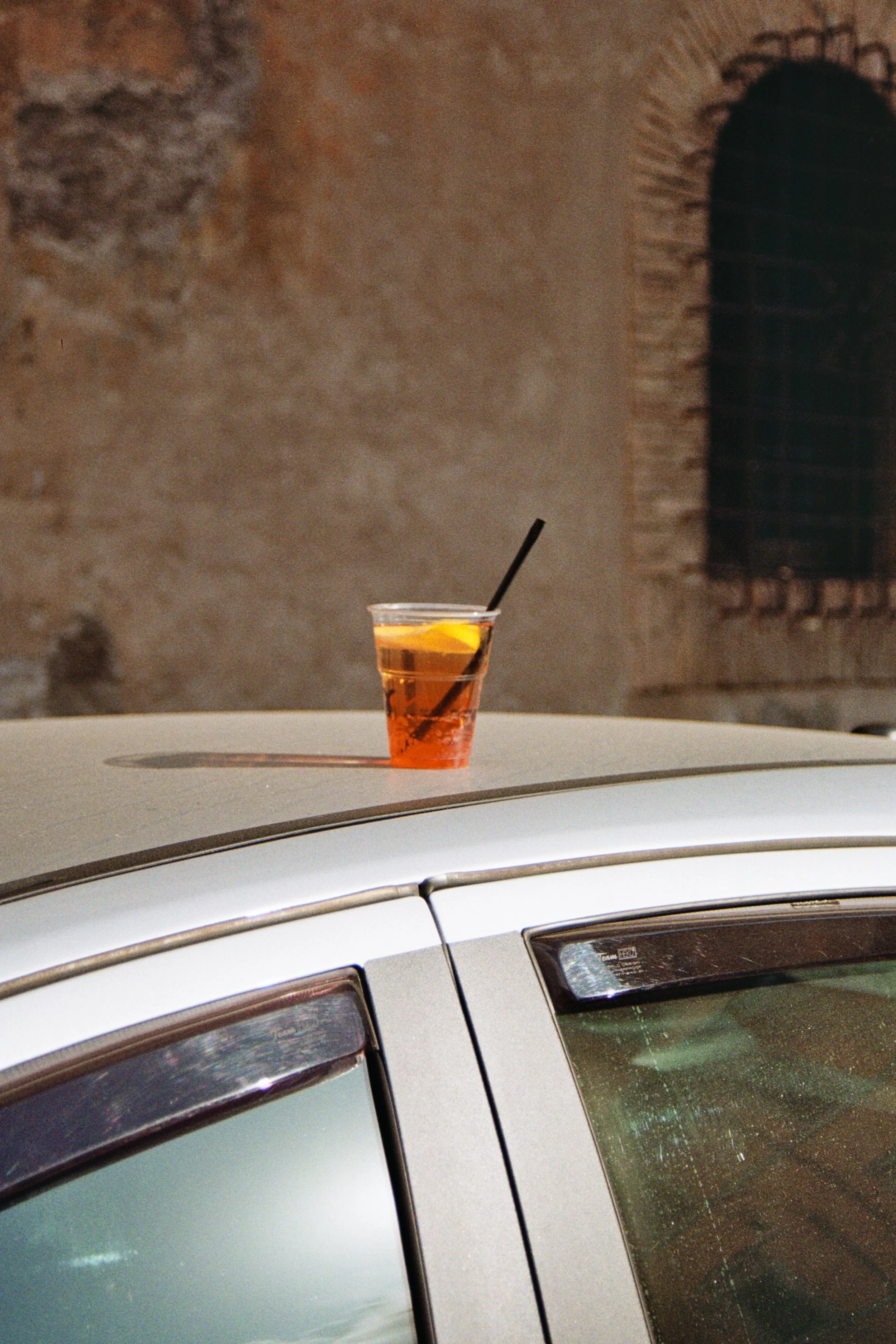 Copo de plástico com uma bebida âmbar, limão e canudo, colocado sobre o teto de um carro, com uma parede de alvenaria ao fundo.