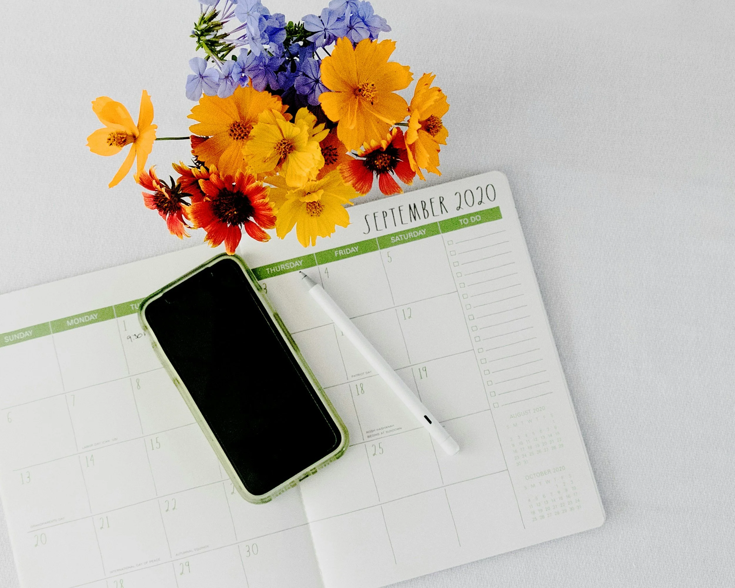 A flat lay of a September 2020 planner, a smartphone in a green case, a white pen, and a bouquet of colorful flowers with purple, yellow, orange, and red blooms on a white surface.