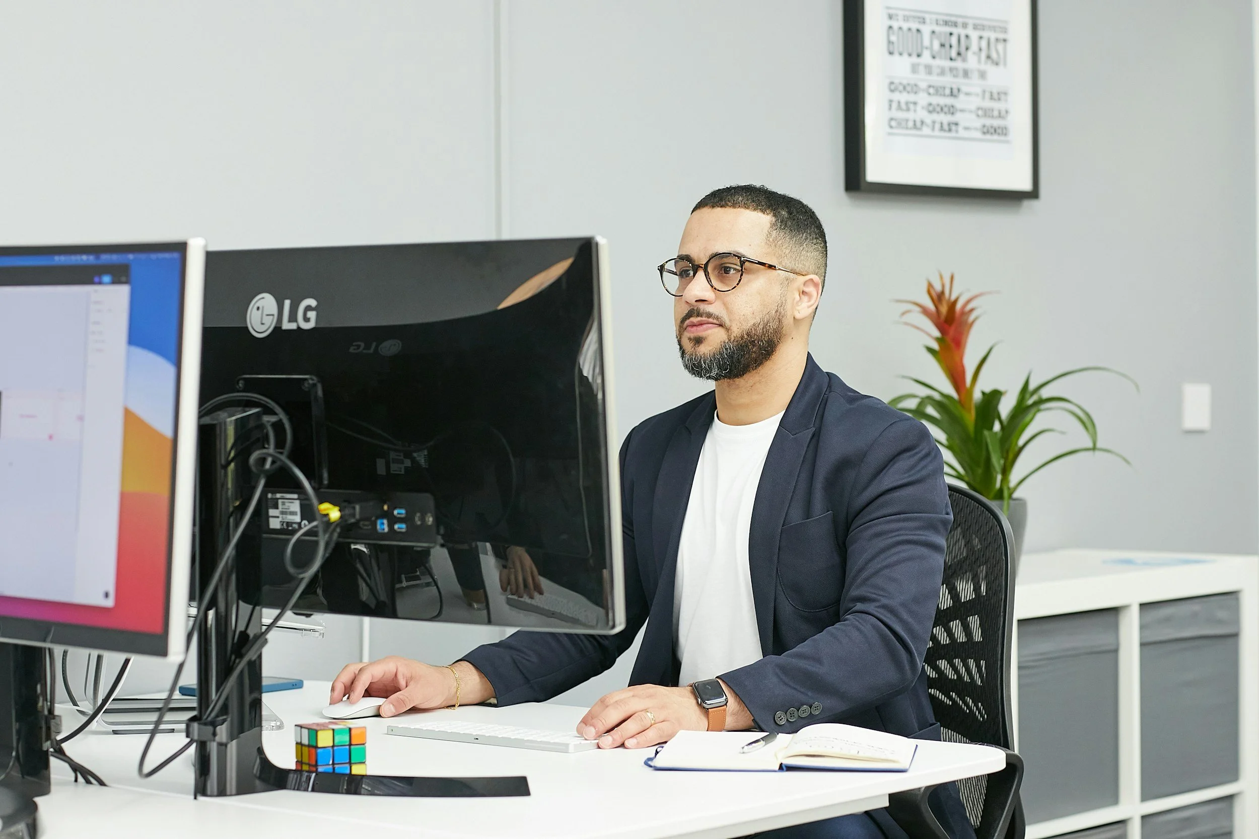 A man with glasses and a beard, wearing a navy blazer, sitting at a desk working on a computer in an office with a white wall, a framed poster, a green and red potted plant, an open notebook, and a Rubik's cube.