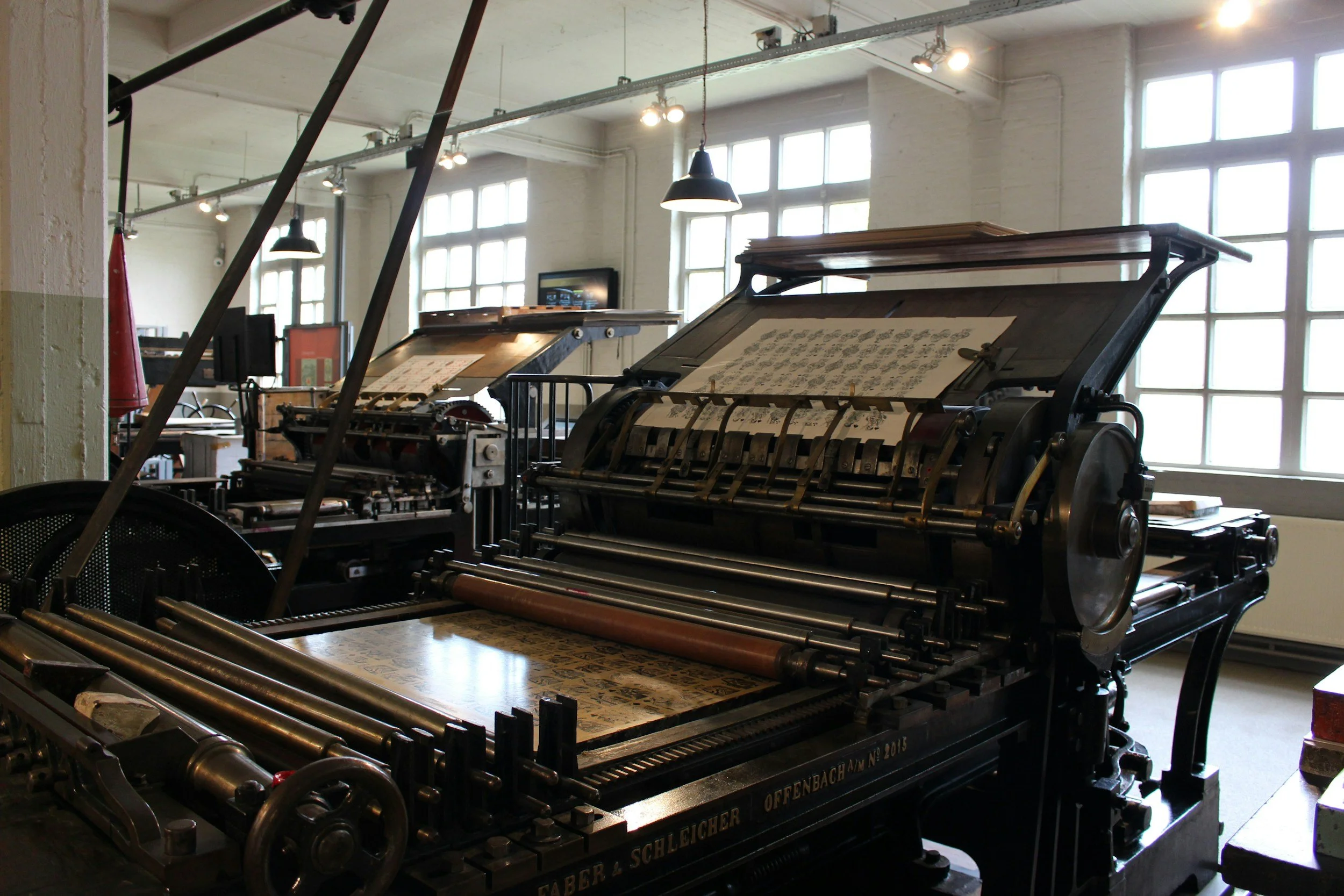 Vintage printing presses inside a well-lit room with large windows and hanging lights.
