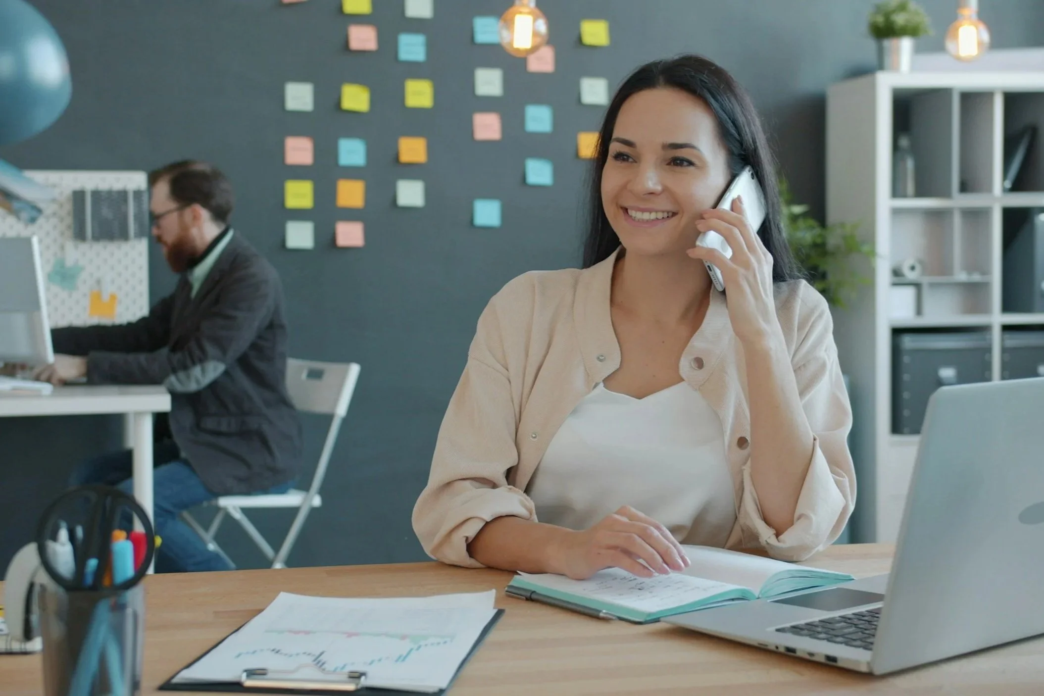 A woman sitting at a desk in an office, smiling, talking on a phone, with a laptop, open notebook, and papers, while a man works at a computer in the background.