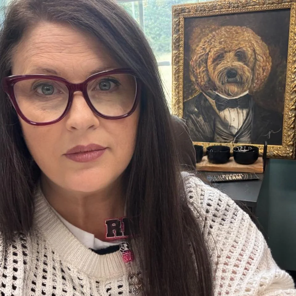A woman with long dark hair and large red glasses takes a selfie in front of a painting of a dog dressed in a tuxedo. The woman wears a white knitted sweater and a necklace with the initials R and S.