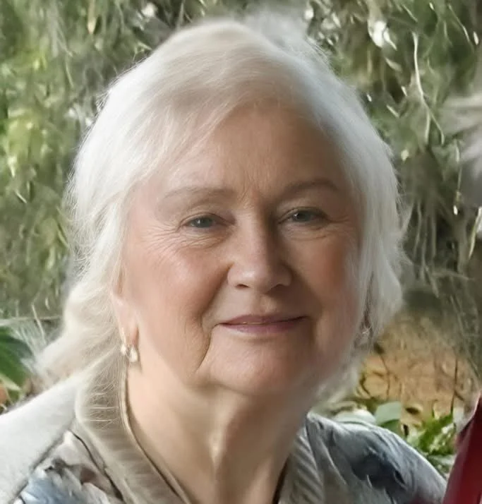 Close-up of an elderly woman with white hair smiling outdoors, blurred green foliage background.