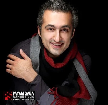 A man smiling, wearing a black jacket, red scarf, and gray overcoat, against a black background.