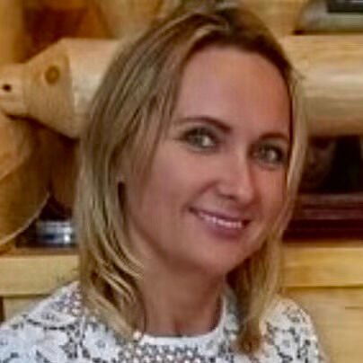 A woman with blonde hair smiling, wearing a white top with lace details, standing indoors near wooden logs.