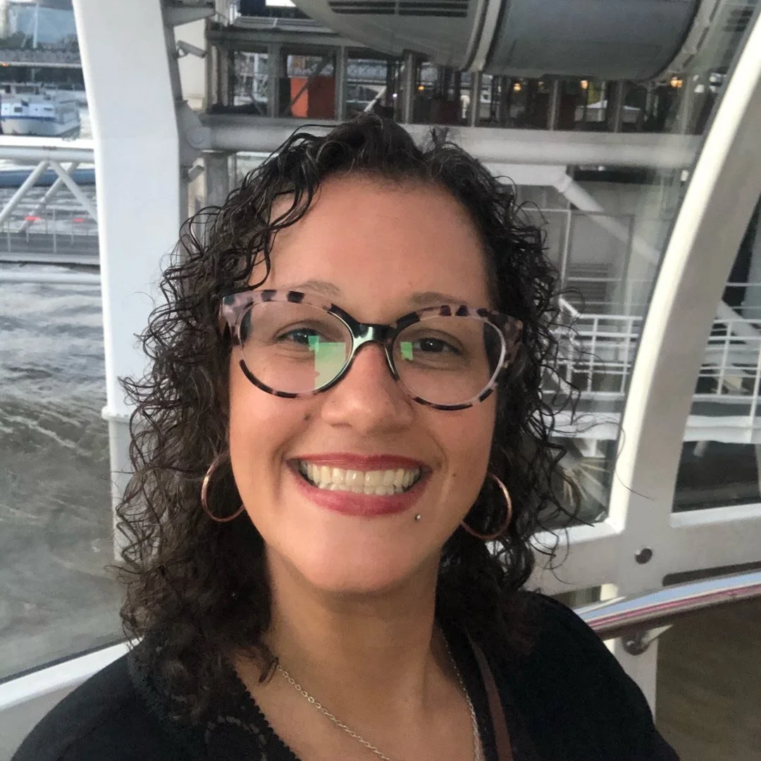 A woman with short curly dark hair, wearing glasses with a tortoiseshell pattern, smiling, in an indoor setting near large windows showing an outdoor dock with boats.