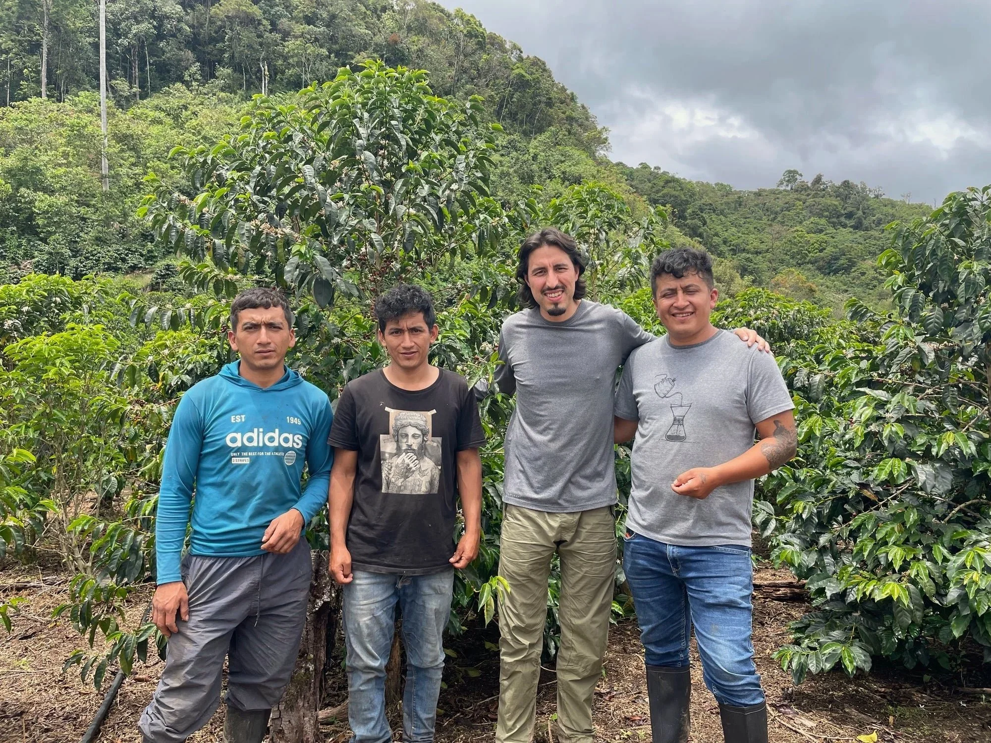 Four men standing together outdoors with lush green trees and a hilly landscape in the background.