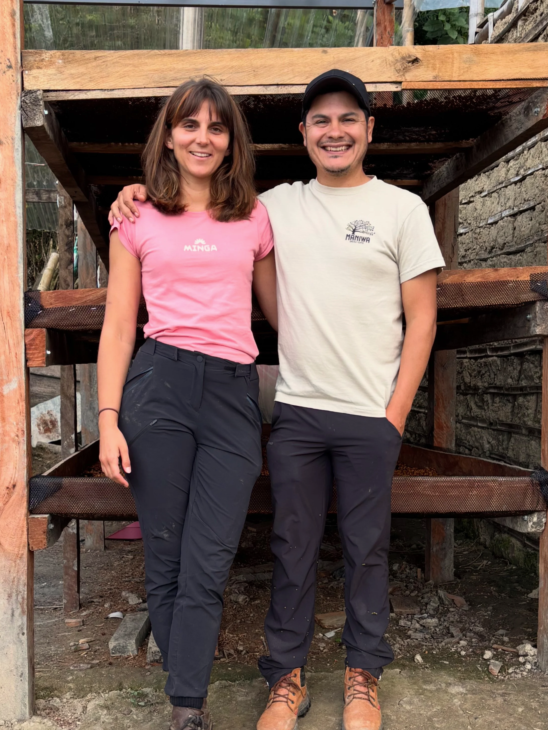 A man and woman standing outdoors in front of a wooden structure, smiling, with greenery in the background, dressed casually in outdoor clothing.