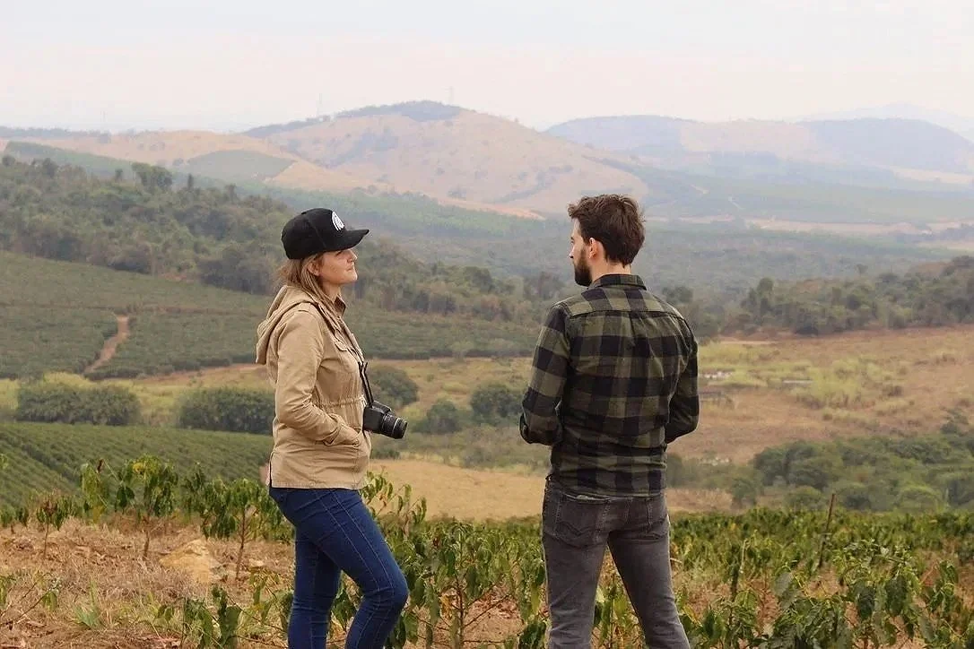 A woman and a man standing outdoors in a rural landscape with rolling hills and farmland in the background. The woman has a camera around her neck and is wearing a beige jacket and black cap. The man has a beard and is wearing a plaid shirt.