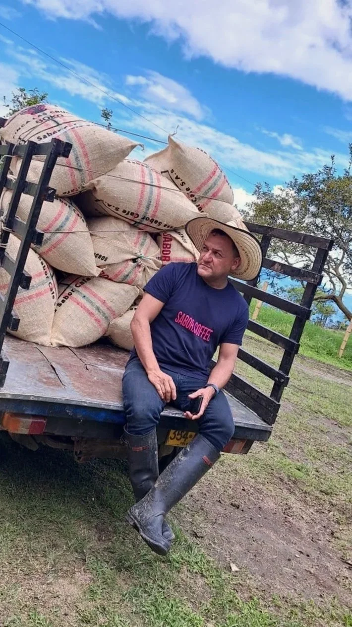 Man sitting on the back of a truck, wearing a large straw hat, boots, and a blue t-shirt with red text, in a rural outdoor setting with bags of coffee beans in the truck bed and a partly cloudy sky.