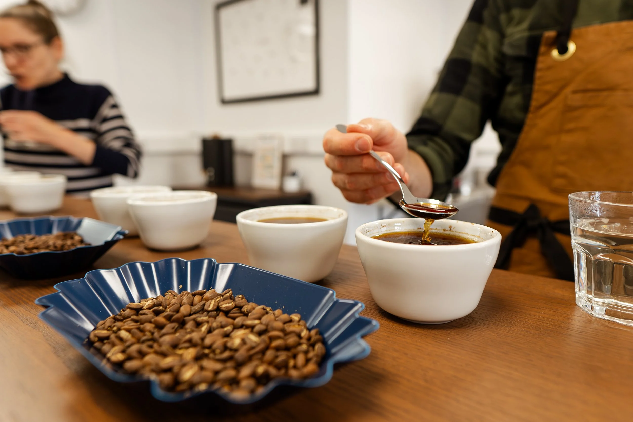 Person at a coffee tasting event with bowls of coffee beans and cups of black coffee on a wooden table.