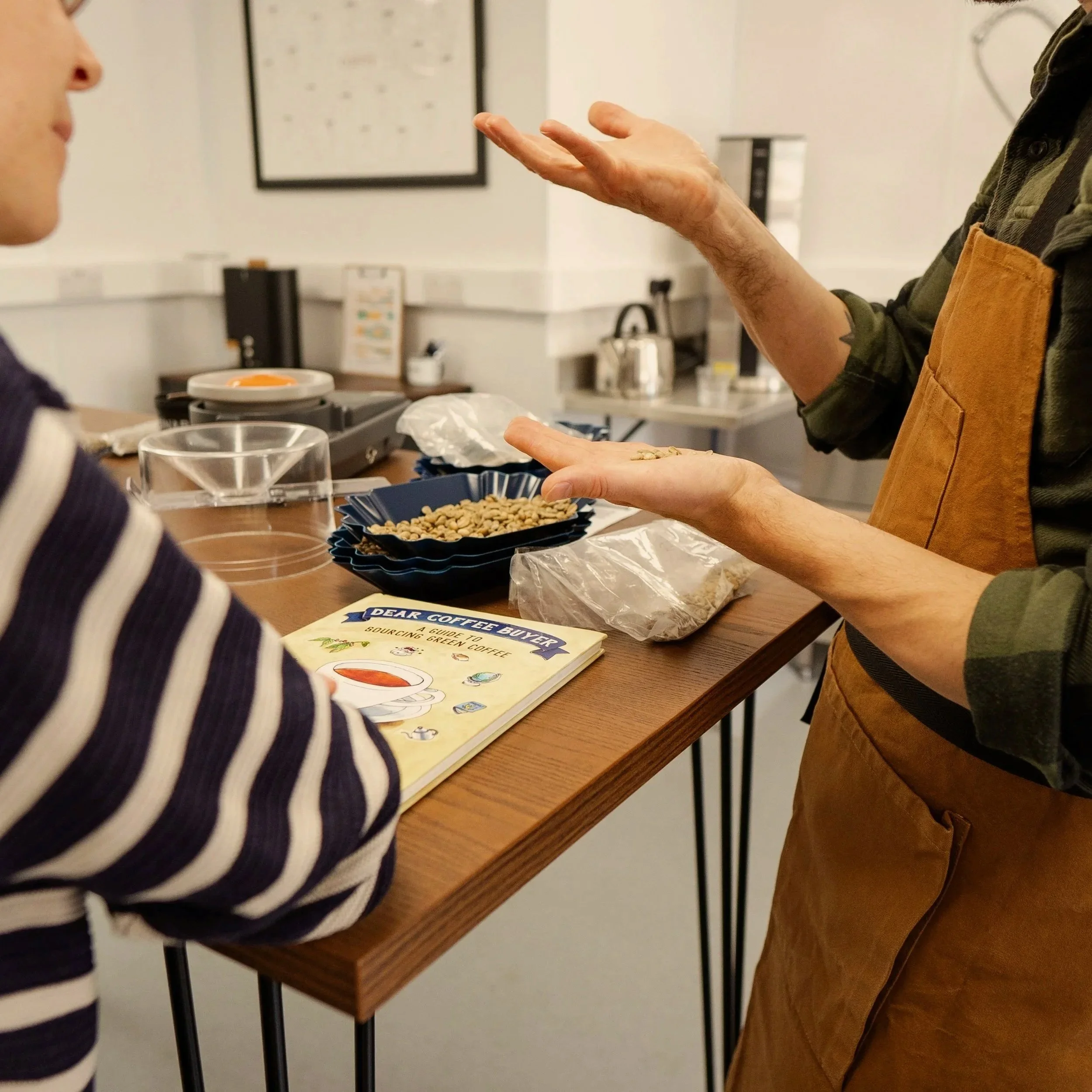 A person in an apron talking to another person at a counter with coffee beans, a book titled 'Dear Coffee Buyer,' and coffee supplies in a lab.
