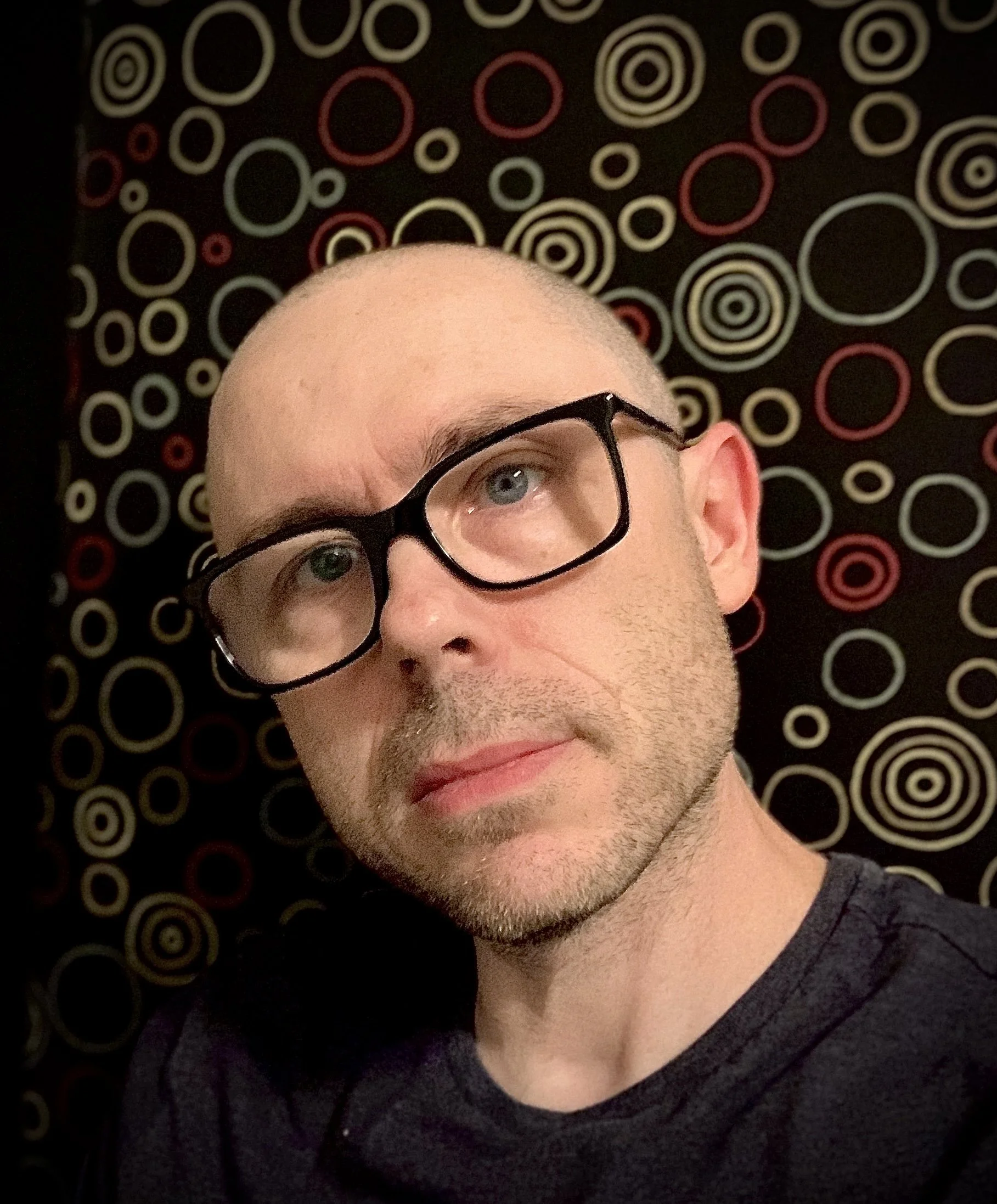 Close-up of a man with glasses, short hair, and stubble, looking slightly upward, against a backdrop of a black wall with colorful circular patterns.