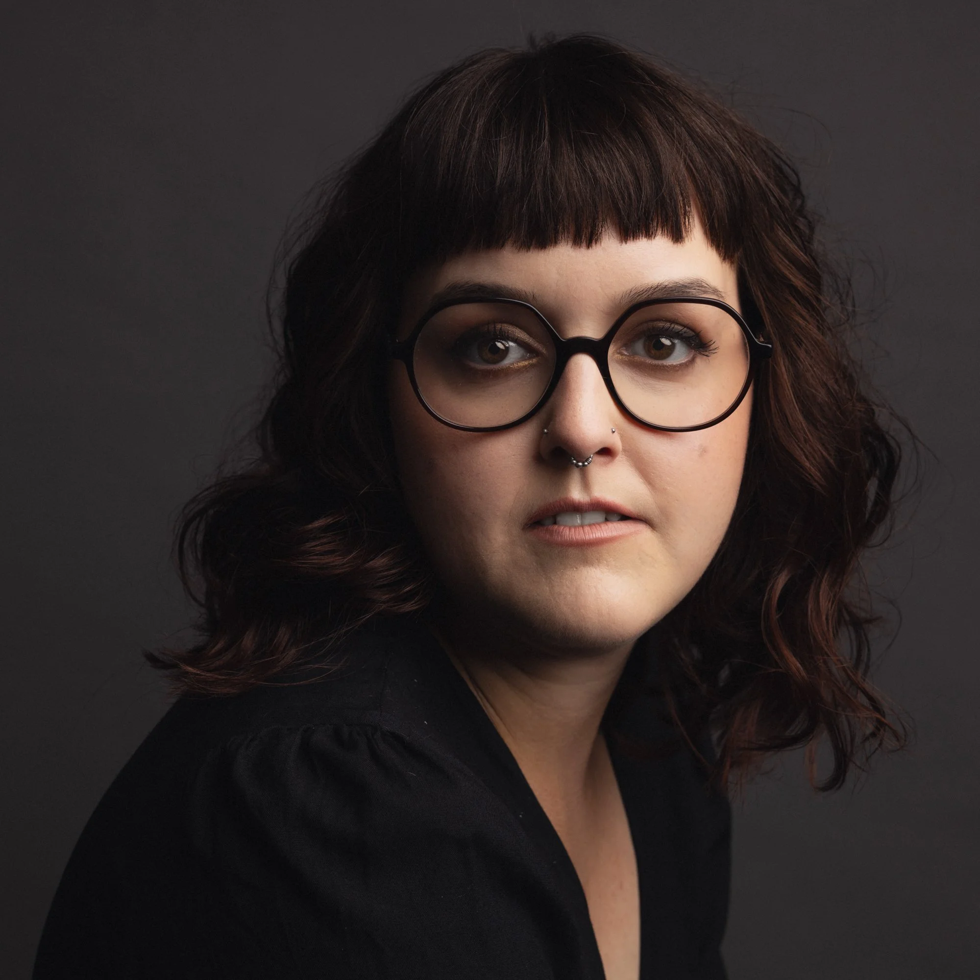 Portrait d'une jeune femme aux cheveux bouclés bruns, portant des lunettes rondes et un piercing au nez, contre un fond sombre.
