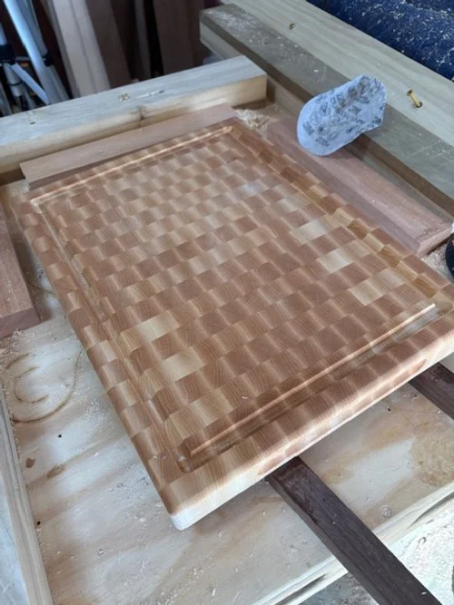 A partially finished wooden cutting board with a checkerboard pattern, resting on a workbench in a woodworking shop.