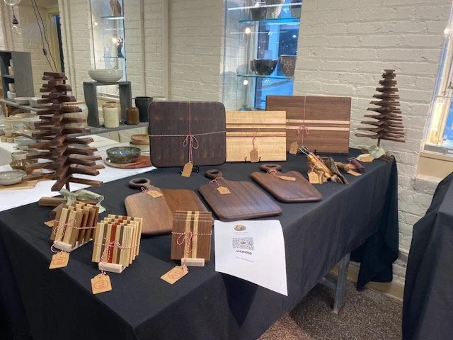 A display table with wooden crafts and gift items, including cutting boards, jewelry, and decorative wooden trees, set against a white brick wall with windows.