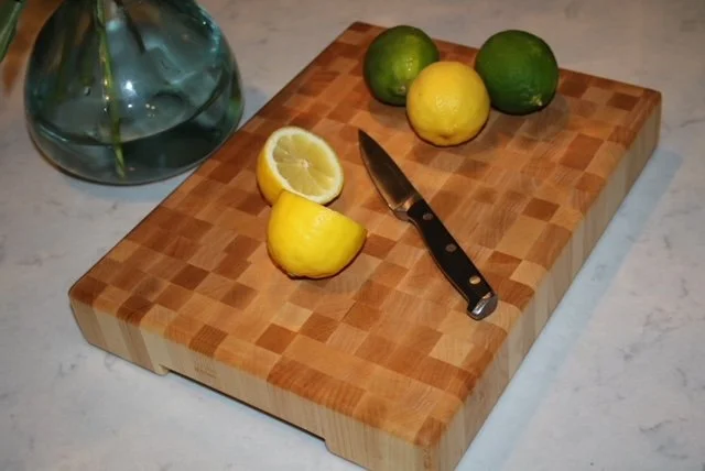 A wooden cutting board with three limes, one lemon, and a lemon half, along with a small kitchen knife. A glass vase or container with water and green leaves is partially visible in the background on a marble countertop.