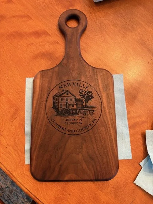 A wooden cutting board with a handle, engraved with a house, tree, and water wheel, and text reading "Newville Cumberland County, PA" along with GPS coordinates.