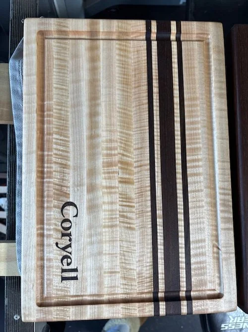 Wooden cutting board with black and dark brown decorative stripes and the text 'Coryell' engraved on the bottom left corner.