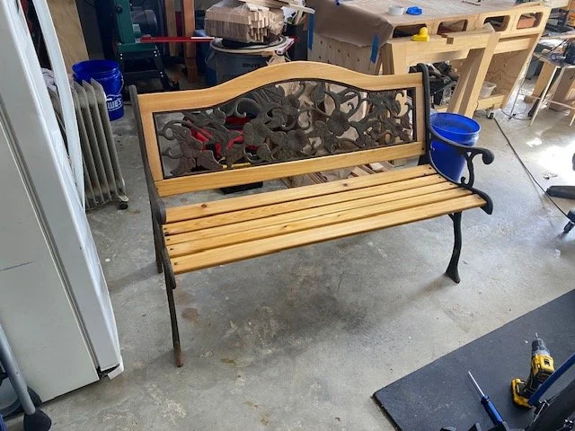 Wooden and metal garden bench in a workshop, with tools and furniture in the background.