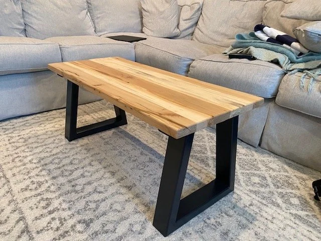 A wooden table with black metal legs in a living room with a gray sofa and folded blankets.