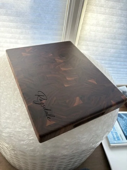 A dark-colored wooden box with a signature that reads "Kayalee" on the top surface, placed on a textured white side table near a window with blinds.