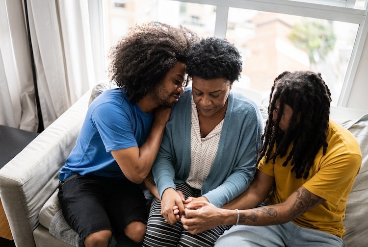 Three women sitting together on a couch in a bright living room, comforting a woman in the middle who appears upset, with two women holding her hands and leaning in support.