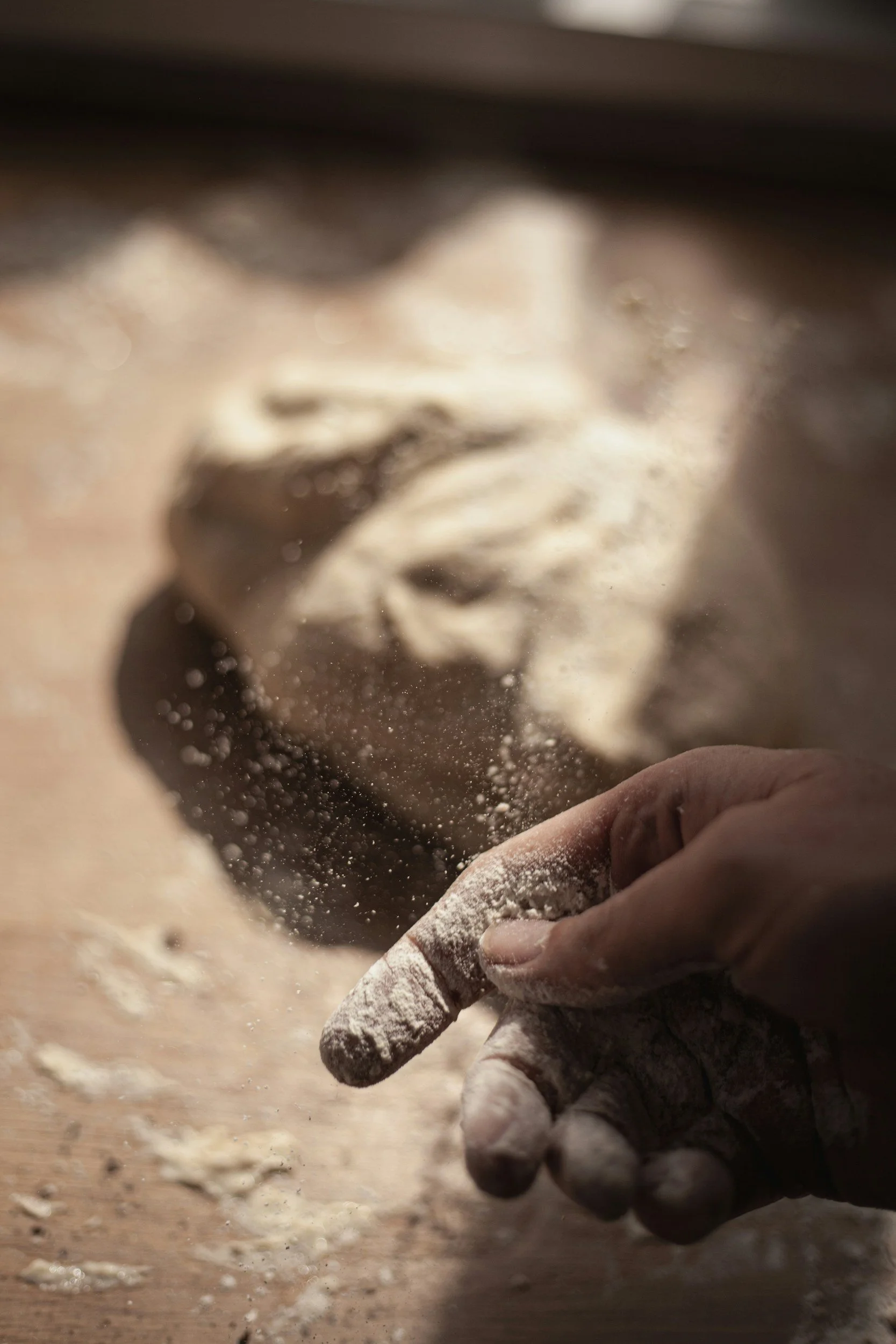 A person's hand dusted with flour, creating a cloud of flour particles as they work with dough on a wooden surface.