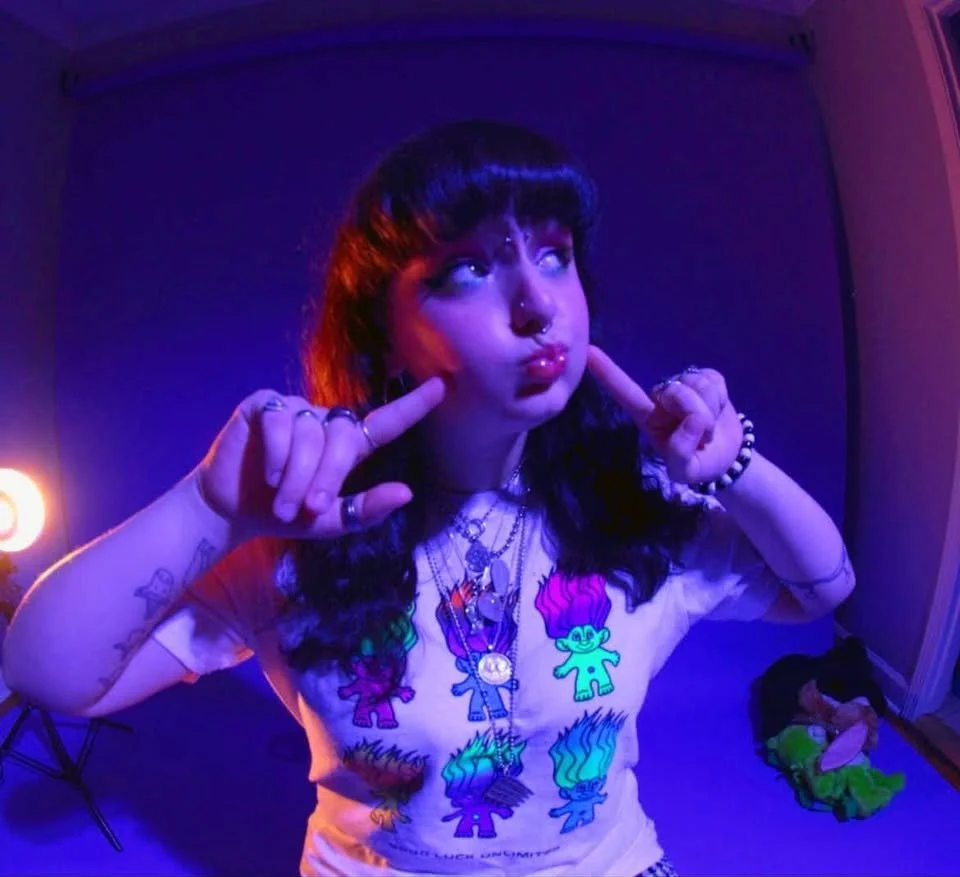 A person with dark hair, multiple necklaces, and face piercings making a playful gesture near their face in a room with purple and pink lighting. They are wearing a colorful T-shirt with cartoon characters.