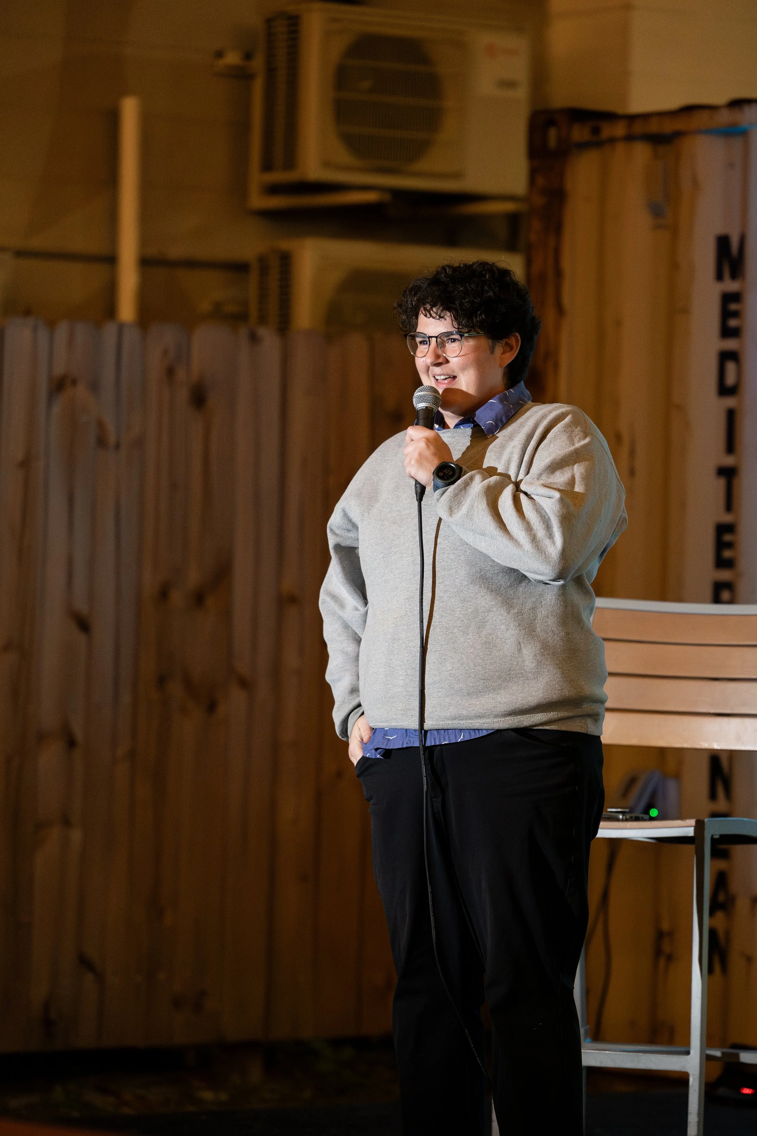 Person speaking into a microphone on stage with a wooden backdrop.
