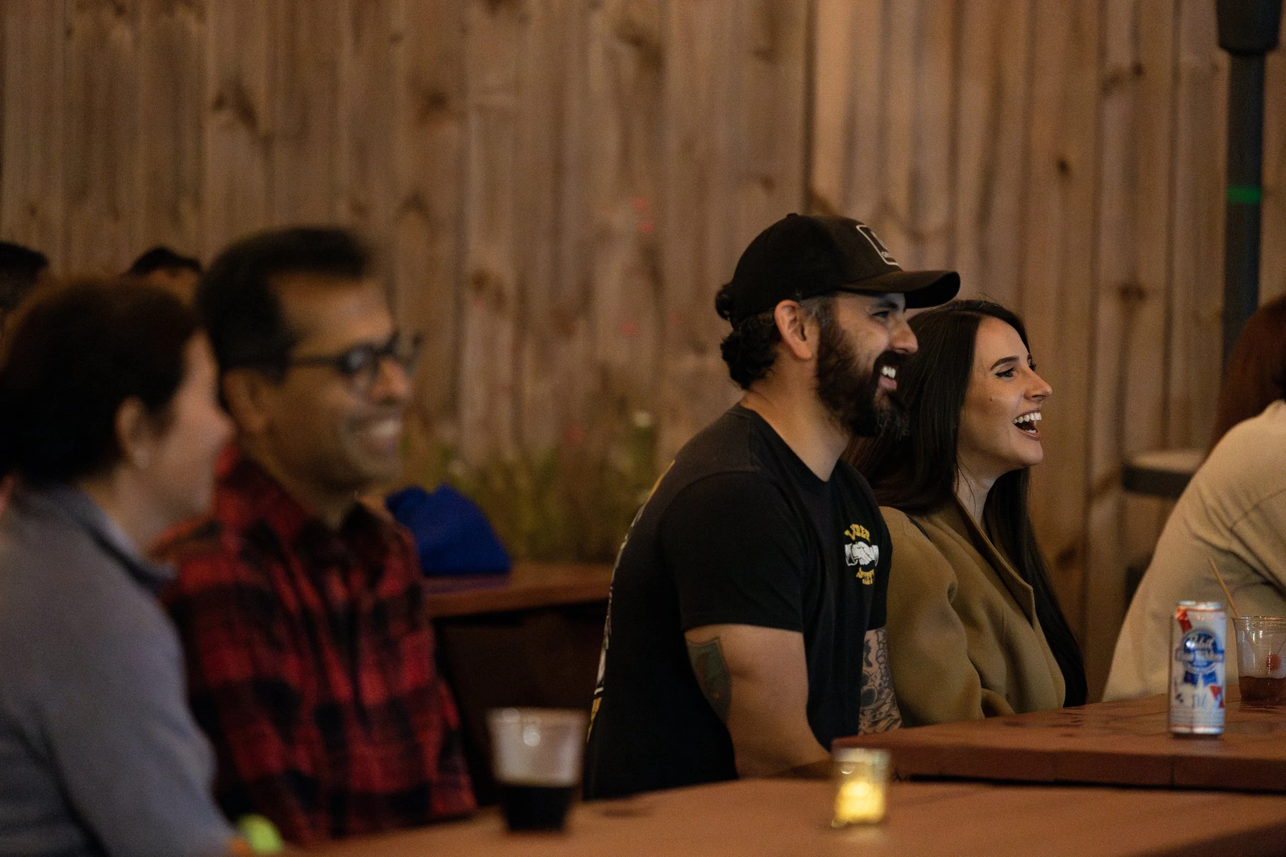 People sitting at a wooden bar or table, smiling and laughing during an event or gathering.