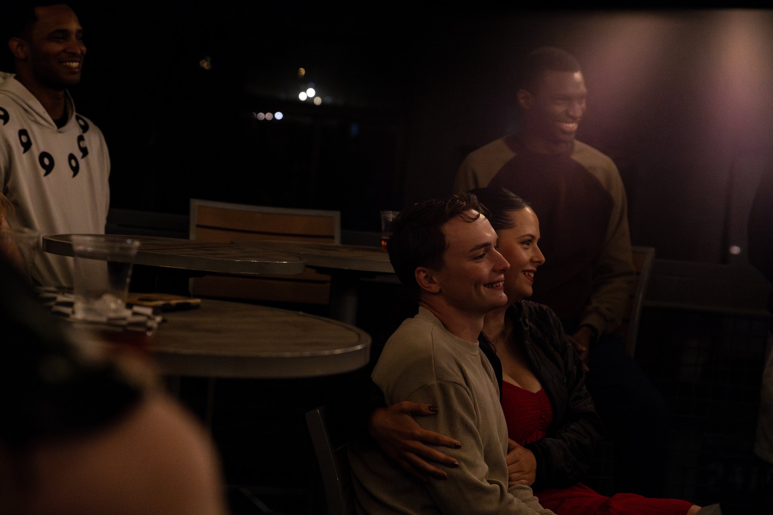 A group of people smiling and enjoying a moment together in a dimly lit indoor setting, with some seated and some standing.
