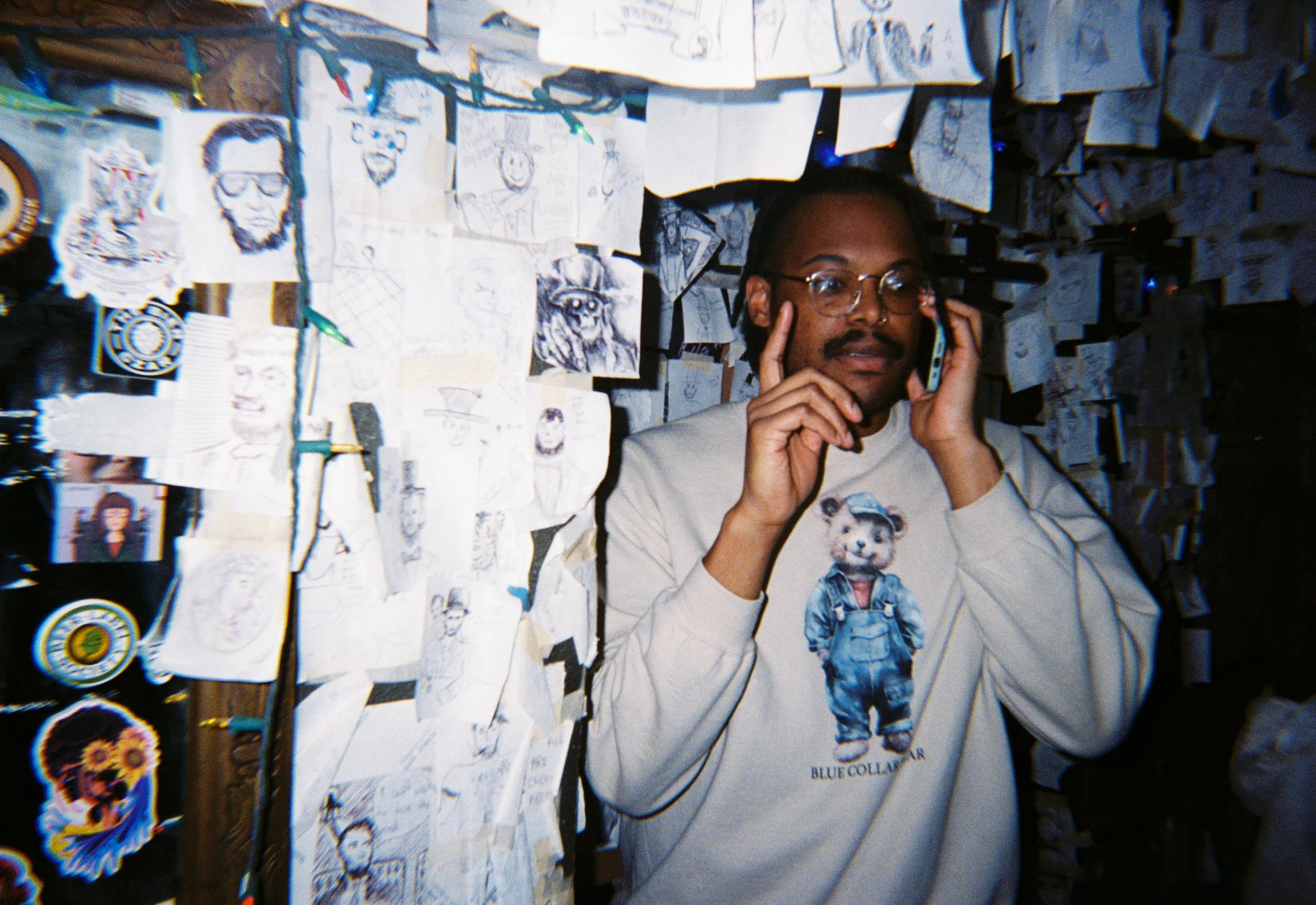 A man with glasses and a beard talking on a cellphone, standing in front of a wall decorated with numerous sketches, drawings, and stickers.