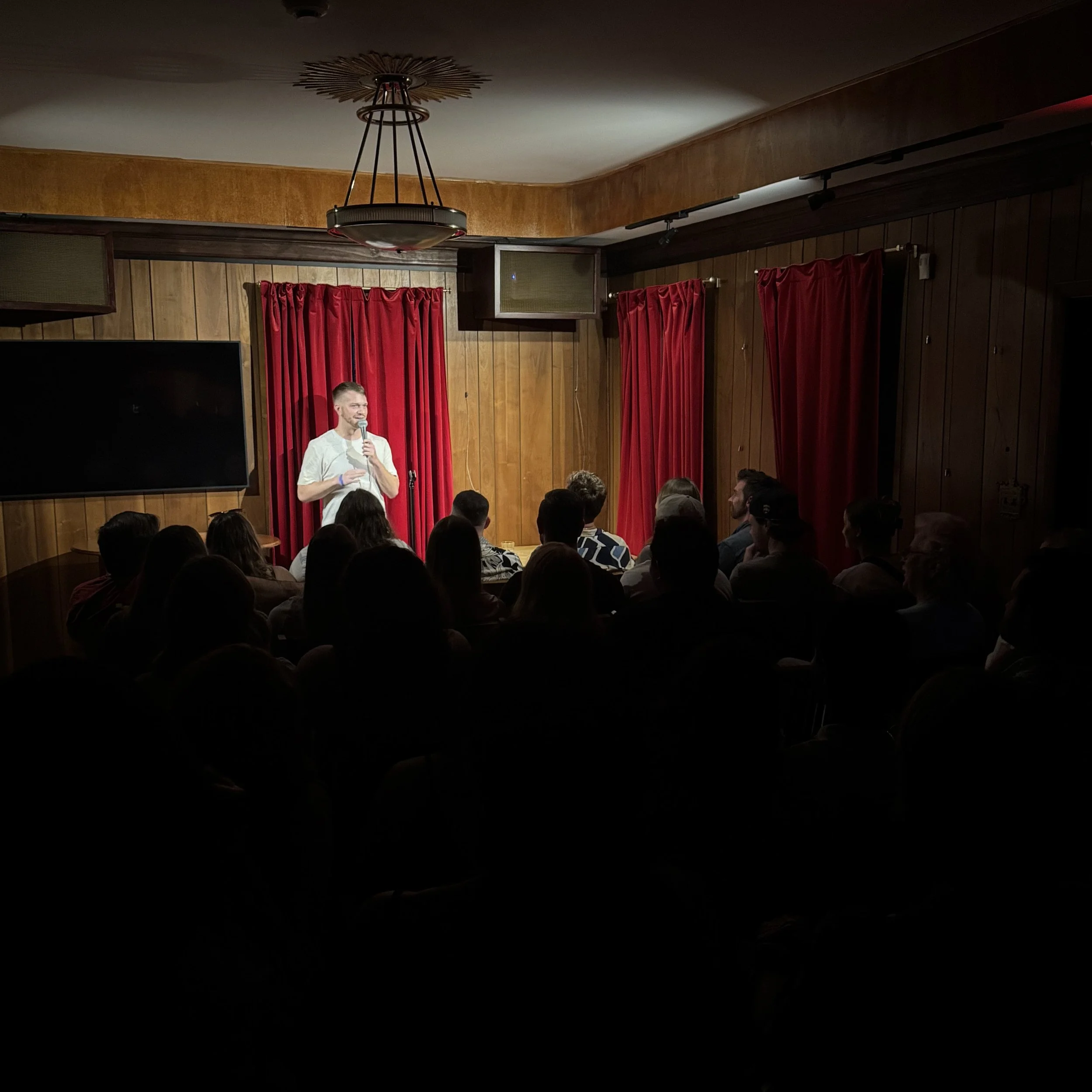A man on stage with a microphone speaking to an audience in a small, wood-paneled comedy club or performance venue. The stage has red curtains and a large black screen on one side.