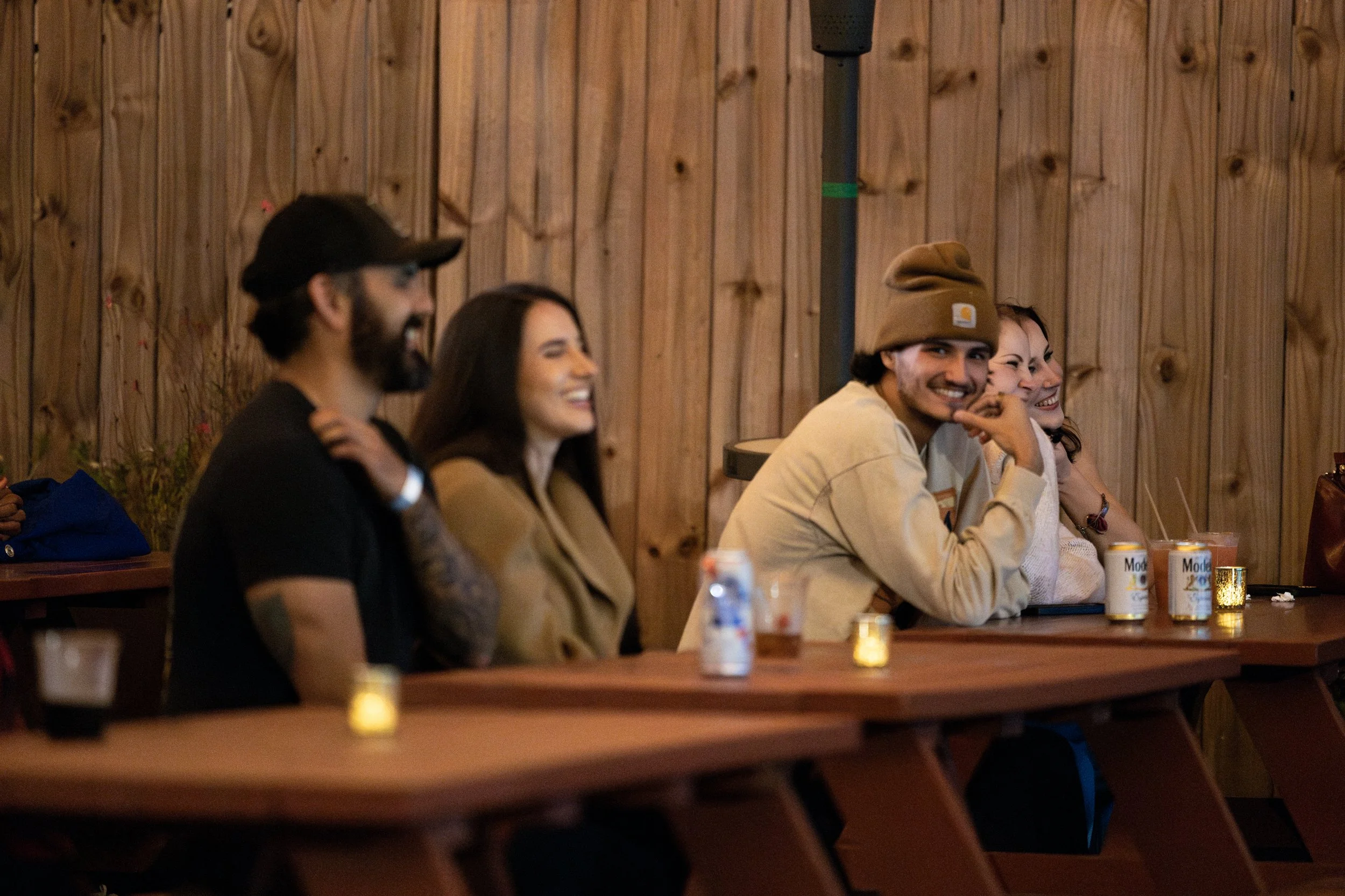 Group of five young adults sitting at a bar or counter, smiling and laughing, with wooden paneling on the walls behind them, drinks in front of them, and small candles on the counter.