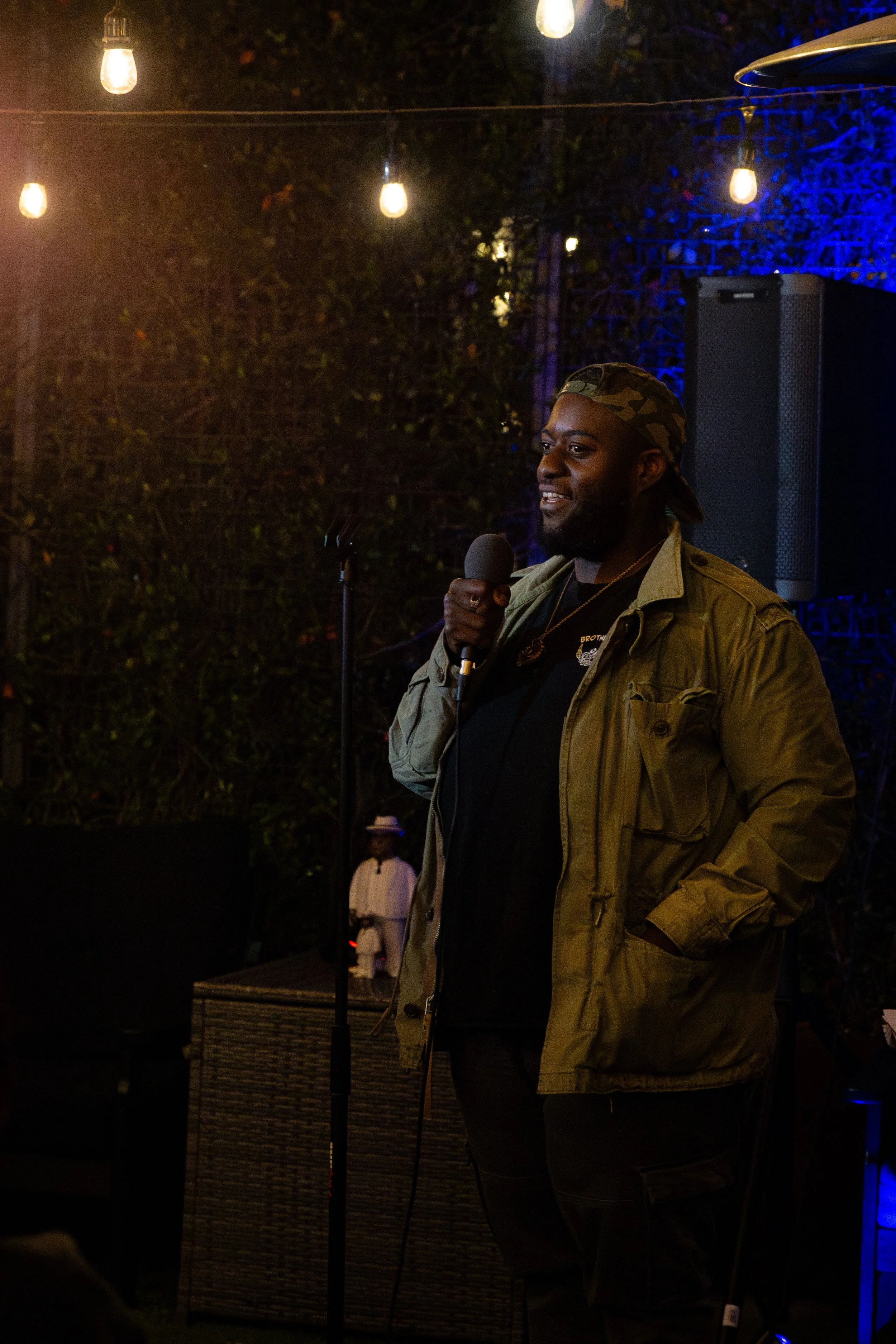 A man with a beard, wearing a camouflage cap and a beige jacket, is holding a microphone and speaking at an outdoor event during the evening. String lights hang above, and there are people and greenery in the background.