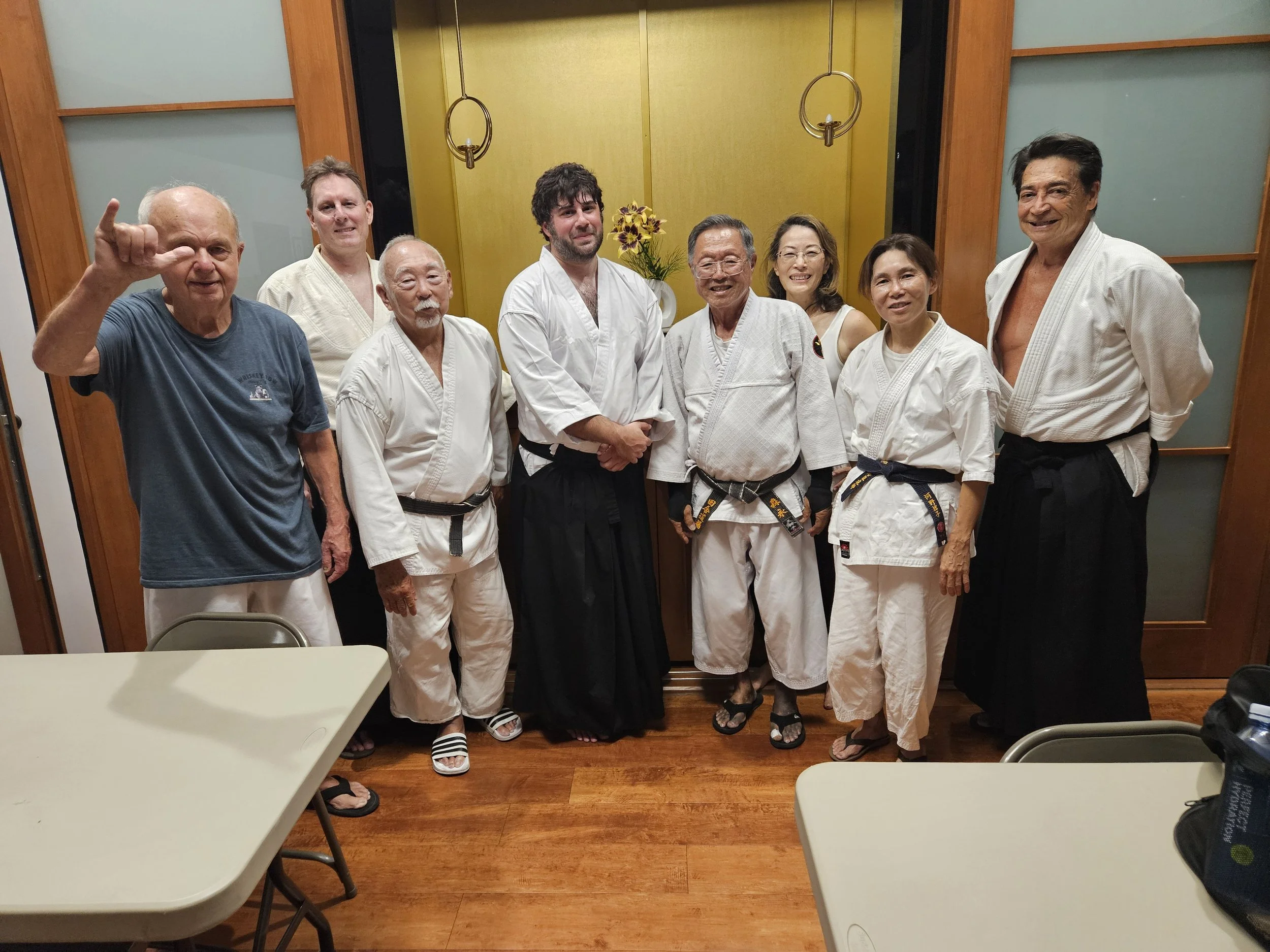 Group of nine people, some in karate uniforms, posing together in a room with wooden floors and a yellow wall.