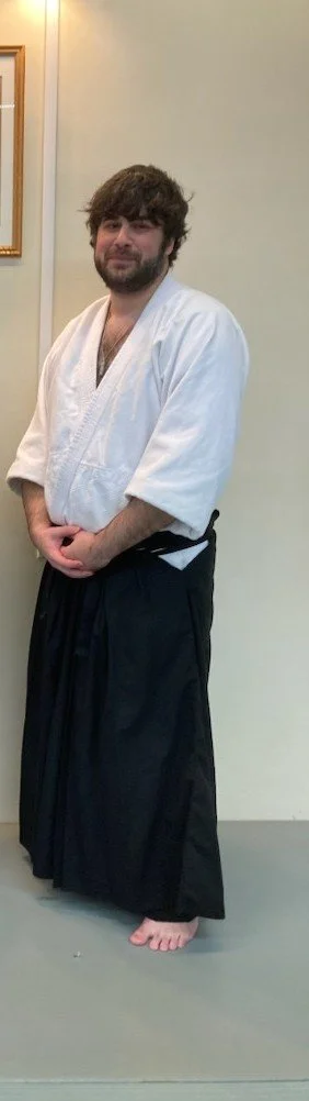 A man wearing a white martial arts gi top and black hakama pants standing barefoot against a beige wall and carpeted floor.